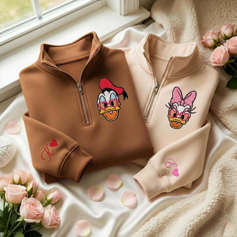 May include: Two quarter-zip sweatshirts, one brown and one beige, featuring embroidered cartoon duck characters wearing sunglasses. The brown sweatshirt has a red heart and the letter "K" embroidered on the sleeve. The beige sweatshirt has a pink heart and the letter "E" embroidered on the sleeve.