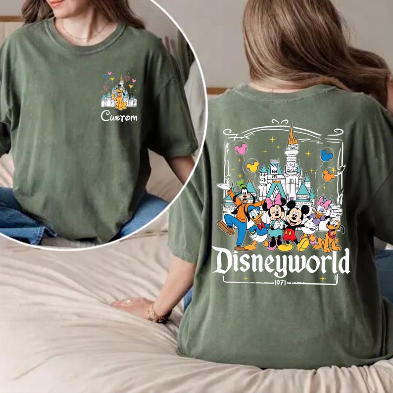 May include: Olive green t-shirt featuring a Disney theme. The front has a small graphic with Pluto and the word "Custom". The back displays a larger design with Mickey, Minnie, Donald, Daisy, Goofy, and the Disney castle, with the text "Disneyworld 1971".