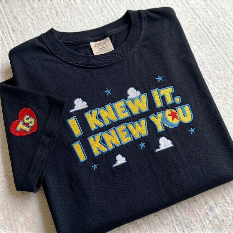 Puede incluir: Una camiseta negra de cuello redondo con texto bordado que dice I KNEW IT, I KNEW YOU en amarillo con bordes azules. El dise&ntilde;o incluye nubes blancas, estrellas azules y una pelota roja con una estrella amarilla. Un parche de coraz&oacute;n rojo con TS est&aacute; en la manga.