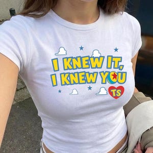 May include: A white short-sleeved t-shirt featuring the yellow text I KNEW IT, I KNEW YOU with a colorful ball icon, a red heart containing the letters TS, and blue cloud and star graphics printed on the front center of the garment.