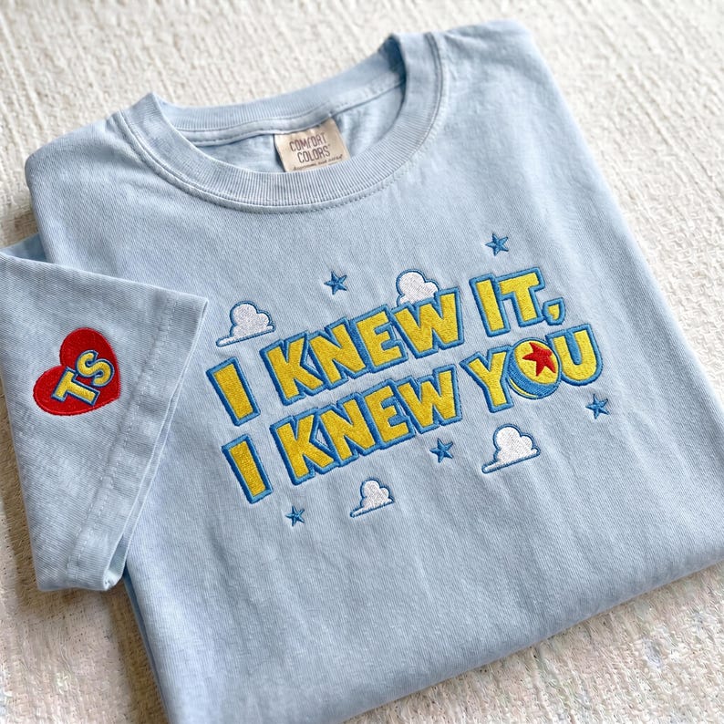 Puede incluir: Una camiseta azul claro con texto bordado que dice I KNEW IT, I KNEW YOU en amarillo con bordes azules. El dise&ntilde;o incluye peque&ntilde;as nubes blancas, estrellas azules y una pelota con una estrella roja. Un coraz&oacute;n rojo con TS est&aacute; en la manga.
