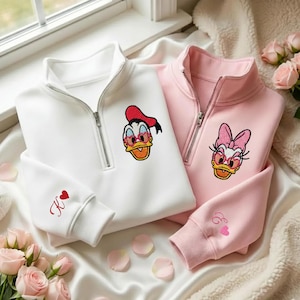 May include: Two zip-up sweatshirts, one white and one pink, featuring embroidered cartoon duck characters wearing heart-shaped sunglasses. The white sweatshirt has a red "K" with a heart on the sleeve. The pink sweatshirt has an "E" with a heart on the sleeve.
