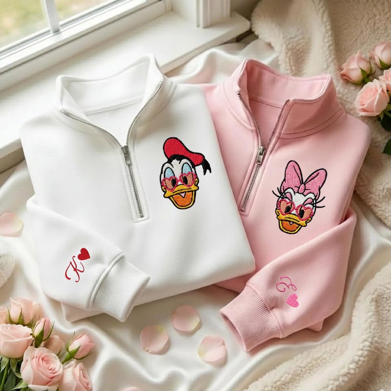 May include: Two zip-up sweatshirts, one white and one pink, featuring embroidered cartoon duck characters wearing heart-shaped sunglasses. The white sweatshirt has a red "K" with a heart on the sleeve. The pink sweatshirt has an "E" with a heart on the sleeve.