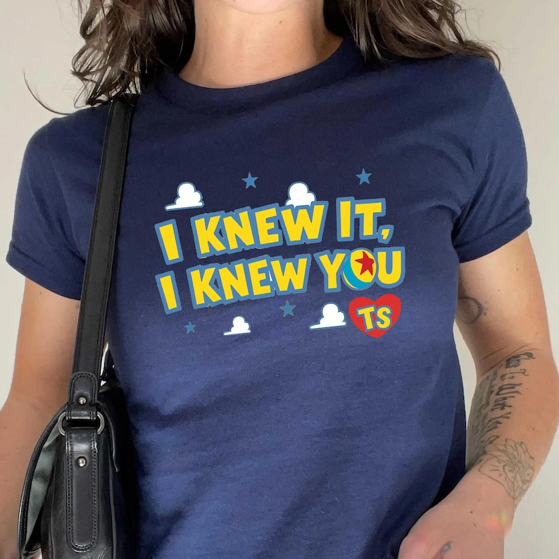 May include: A navy blue short-sleeve t-shirt featuring the yellow text I KNEW IT, I KNEW YOU with a colorful ball icon replacing the letter O, a red heart containing the letters TS, and small white cloud and blue star graphics printed on the front.