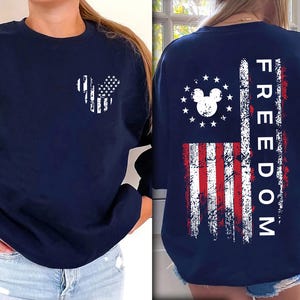 May include: Navy blue sweatshirt with a patriotic Mickey Mouse-shaped American flag design on the front. The back features a distressed American flag with the word "FREEDOM". The design uses red, white, and blue colours.