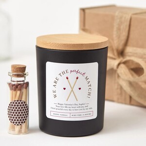 May include: A black candle with a wooden lid and a label that reads "We are the perfect match!" with crossed matchsticks. A small glass bottle of matches and a gift box are also in the image. The candle scent is vanilla and has a burn time of 25 hours.
