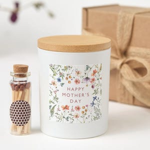 May include: A white candle with a wooden lid and a floral design label that reads "Happy Mother's Day." A small glass bottle of matches is next to the candle. A wrapped gift box is in the background.