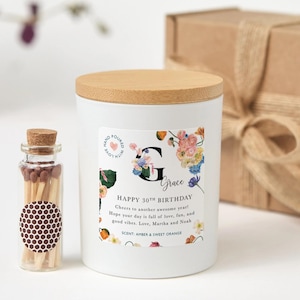 Könnte beinhalten: Eine weiße Kerze mit Holzdeckel und floralem Design, beschriftet mit "Happy 30th Birthday Grace". Neben der Kerze steht eine kleine Glasflasche mit Streichhölzern. Der Duft der Kerze ist Amber & Sweet Orange.