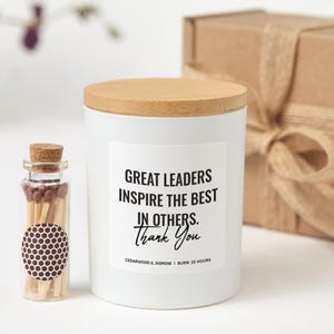 Può includere: Una candela bianca con coperchio in legno e la scritta "GREAT LEADERS INSPIRE THE BEST IN OTHERS. Thank You." Accanto alla candela c'è un piccolo flacone di vetro con fiammiferi. Sullo sfondo c'è una confezione regalo.