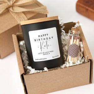 Könnte beinhalten: Eine schwarze Kerze mit Holzdeckel und dem Aufdruck "Happy Birthday Kate". Die Kerze befindet sich in einem Karton mit Papier und einer kleinen Glasflasche mit Streichhölzern. Duft: Zedernholz & Jasmin.