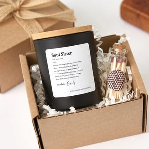 Puede incluir: Una vela negra con tapa de madera y una etiqueta que dice "Soul Sister" en una caja de regalo con una pequeña botella de vidrio de cerillas. La etiqueta de la vela incluye una definición y la fragancia es ámbar y naranja dulce.