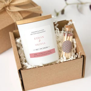 Puede incluir: Una vela blanca con tapa de madera y una etiqueta que dice "Felicitaciones por tu compromiso" con los nombres Ethan & Olivia y la fecha 02/19/2026. La vela está en una caja de cartón con un pequeño frasco de vidrio con cerillas.