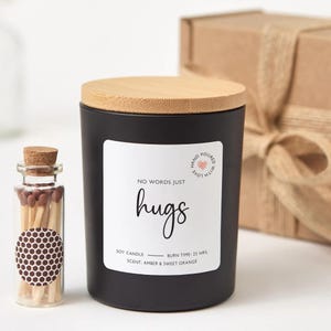 May include: A black soy candle with a wooden lid and a label that reads "hugs." A small glass bottle of matches is next to the candle. The candle's scent is amber and sweet orange.