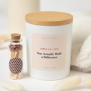 May include: A white candle in a cylindrical container with a light wood lid and a label reading Sparks & More, Smells Like You Actually Made a Difference, Vegan Soy Wax. Beside it is a small glass bottle with a cork stopper containing brown matches.