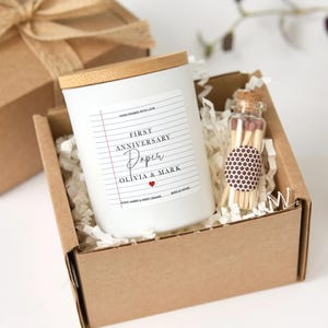 Könnte beinhalten: Eine weiße Kerze mit Holzdeckel und dem Aufdruck "FIRST ANNIVERSARY Paper OLIVIA & MARK" befindet sich in einer Pappschachtel mit einer kleinen Glasflasche Streichhölzer. Die Schachtel ist mit weißen Papierschnipseln gefüllt und mit einem braunen Band gebunden.