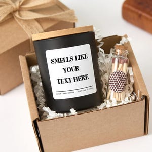 May include: A black candle with a wooden lid and a white label that reads "Smells Like Your Text Here." The candle is in a cardboard box with a small glass bottle of matches. The candle scent is amber and sweet orange.