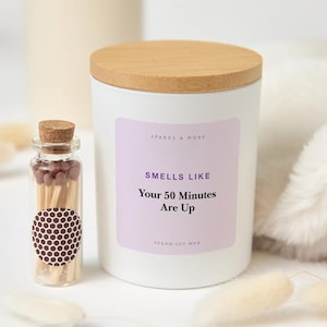 May include: A white candle in a cylindrical container with a light wooden lid and a light purple label that reads Sparks & More, Smells Like Your 50 Minutes Are Up, Vegan Soy Wax. Beside it is a small glass bottle filled with brown-tipped matches.