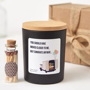 May include: A black candle in a jar with a bamboo lid, featuring a label that reads: YOU SHOULD HAVE MOVED CLOSER TO ME, BUT CONGRATS ANYWAY... with an illustration of a moving van and balloons. Beside it is a small glass bottle filled with matches.