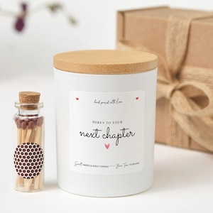 Puede incluir: Una vela blanca con tapa de madera y la inscripción "Here's to your next chapter". Junto a la vela hay un pequeño frasco de vidrio con cerillas. La vela tiene aroma a ámbar y naranja dulce y tiene un tiempo de combustión de 25 horas.