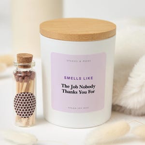 May include: A white candle in a cylindrical container with a light wood lid and a label that reads Sparks & More, Smells Like The Job Nobody Thanks You For, Vegan Soy Wax. Next to it is a small glass bottle filled with matches and a honeycomb sticker.