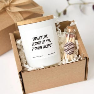 May include: A white candle with a wooden lid and the text "SMELLS LIKE GEORGE HIT THE F*CKING JACKPOT" sits in a cardboard box with shredded paper. A small glass bottle of matches is also in the box. The candle is labeled "CEDARWOOD & JASMINE" and "BURN: 25 HOURS".