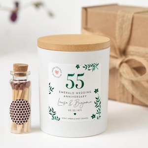 May include: A white candle with a wooden lid and a label that reads "55 Emerald Wedding Anniversary." The label includes the names "Louisa & Benjamin" and the date "05.23.1971." A small glass bottle of matches is next to the candle.