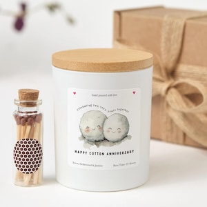 以下が含まれることがあります： 木製の蓋が付いた白いキャンドルで、「Happy Cotton Anniversary」と書かれたラベルが付いています。ラベルには2つの漫画の綿球が描かれています。キャンドルの隣には、マッチが入った小さなガラス瓶があります。キャンドルの香りはシダーウッド＆ジャスミンです。