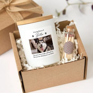 May include: A white candle with a bamboo lid, a photo of a dog, and the text "In Loving Memory of Bella" in a cardboard box with matches. The text below the photo reads "Your paws may no longer walk beside us. But your love will always be in our hearts."