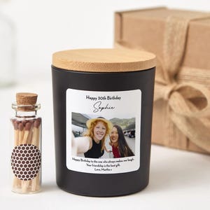 Könnte beinhalten: Eine schwarze Kerze mit Holzdeckel und einem Etikett mit der Aufschrift "Happy 30th Birthday Sophie". Neben der Kerze steht eine kleine Glasflasche mit Streichhölzern. Im Hintergrund befindet sich eine Geschenkbox.