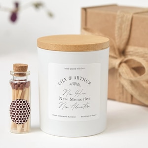 Puede incluir: Una vela blanca con tapa de madera y una etiqueta que dice "Lily & Arthur, New Home, New Memories, New Adventure". Una pequeña botella de cristal con cerillas está al lado de la vela. La fragancia de la vela es Cedarwood & Jasmine.