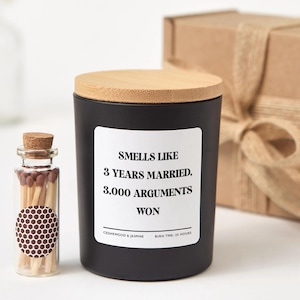 Könnte beinhalten: Eine schwarze Kerze mit Holzdeckel und einem Etikett mit der Aufschrift "Smells Like 3 Years Married, 3,000 Arguments Won". Neben der Kerze steht eine kleine Glasflasche mit Streichhölzern. Die Kerze ist nach Zedernholz und Jasmin duftend.