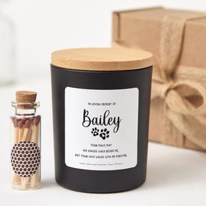 May include: A black candle with a wooden lid and a label that reads "In Loving Memory of Bailey." A small glass bottle of matches is next to the candle. A brown gift box is in the background.