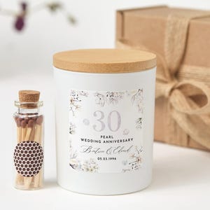 Könnte beinhalten: Eine weiße Kerze mit Holzdeckel und einem Etikett mit der Aufschrift "30 Perlenhochzeitstag". Neben der Kerze steht eine kleine Glasflasche mit Streichhölzern. Im Hintergrund befindet sich eine Geschenkbox mit Schleife.