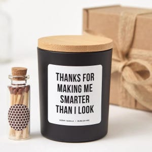 May include: A matte black candle jar with a bamboo lid and a white label reading THANKS FOR MAKING ME SMARTER THAN I LOOK, SCENT: VANILLA | BURN: 25 HRS. Next to it is a small glass bottle with a cork stopper containing wooden matches with brown tips.