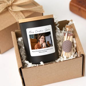 以下が含まれることがあります： 木製の蓋と写真とテキストが付いたラベルが付いた黒いキャンドル。キャンドルは、細断された紙が入った段ボール箱の中にあり、小さなガラス瓶のマッチの隣にあります。ラベルのテキストは「メリークリスマス、ソフィー」と書かれています。