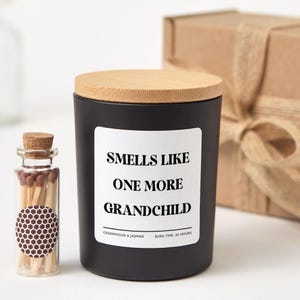 May include: A black candle with a wooden lid and a label that reads "Smells Like One More Grandchild." A small glass bottle of matches is next to the candle. The candle is cedarwood and jasmine scented and has a 25-hour burn time.
