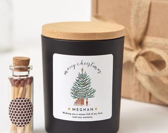 Könnte beinhalten: Eine schwarze Kerze mit Holzdeckel und einem Etikett mit der Aufschrift "merry christmas" und einer Weihnachtsbaumillustration. Neben der Kerze steht eine kleine Glasflasche mit Streichhölzern. Der Text "MEGHAN" steht ebenfalls auf dem Etikett.