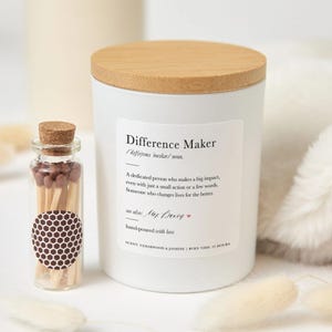 Può includere: Una candela bianca con coperchio in legno e un'etichetta con la scritta "Difference Maker". Accanto alla candela c'è una piccola bottiglia di vetro con tappo di sughero e fiammiferi. La candela profuma di legno di cedro e gelsomino e dura 25 ore.