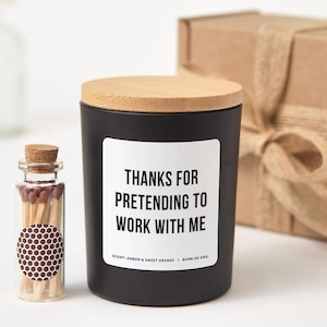 Puede incluir: Una vela negra con tapa de madera y una etiqueta que dice "THANKS FOR PRETENDING TO WORK WITH ME". Una pequeña botella de cristal con cerillas está junto a la vela. La vela tiene aroma a ámbar y naranja dulce, con 25 horas de duración.