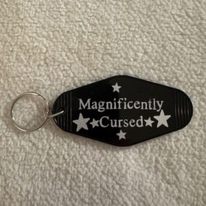 May include: Black keychain tag with the words "Magnificently Cursed" in white, with star accents. The tag has a retro motel key design and a silver key ring. The background is a white, textured surface.