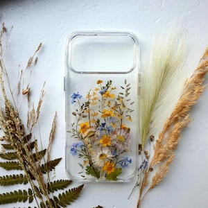 May include: Clear phone case with pressed flowers. The case features a variety of colorful dried flowers, including yellow, blue, and white blooms. The case is surrounded by dried grasses and ferns.