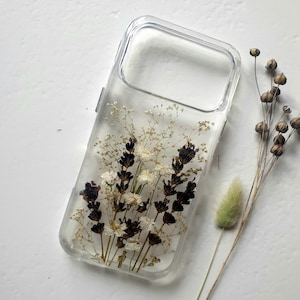 Puede incluir: Funda transparente para teléfono con flores secas incrustadas, incluyendo lavanda y flores blancas. La funda tiene forma rectangular con esquinas redondeadas. Una ramita de flores secas y una planta verde y esponjosa están al lado.