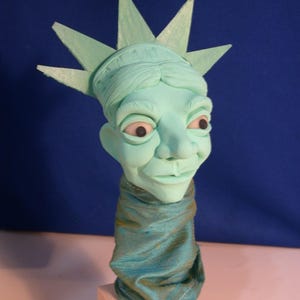May include: A light green sculpture of a head wearing a spiked crown and a teal fabric neck covering. The sculpture is mounted on a square wooden block with the text LADY LIBERTY written in black ink on the front face of the wooden base.