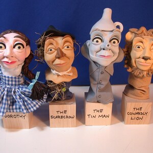 May include: Four character busts from The Wizard of Oz: Dorothy in a blue gingham dress, the Scarecrow with a hat, the Tin Man in a gray suit, and the Cowardly Lion with a mane. Each character is mounted on a wooden block with their name.