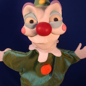 May include: A handmade clown puppet with a white face, red nose, and blue-rimmed eyes. The puppet wears a green satin outfit with a white collar and an orange felt button. A green party hat with colorful pom-poms completes the look.