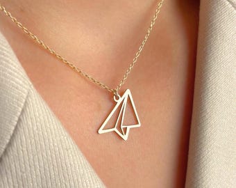 May include: A gold-colored necklace featuring a paper airplane pendant. The pendant is a minimalist design, with clean lines forming the shape of a paper airplane. The necklace is worn against a neutral-toned blazer.