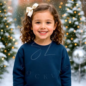 Può includere: Una bambina sorridente con capelli castani ricci e un fiocco color crema. Indossa una felpa blu navy con il testo "MOCKUPS" e "SL". Lo sfondo presenta una scena innevata con alberi di Natale e luci scintillanti.