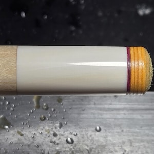 May include: Close-up of a pool cue tip. The tip is a light tan color with a dark purple accent ring. The cue shaft is white with a light tan wooden base.
