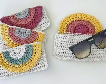 May include: Handmade crochet pouches in various colors, featuring a rainbow design. One pouch holds sunglasses. The pouches are white with a rainbow design in shades of pink, yellow, gray, and teal. The sunglasses have black frames.