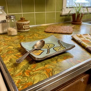 May include: A square, light blue ceramic dish with curved edges featuring a hand-painted design of three stylized leaves in yellow and green. A silver-toned metal spoon rests diagonally across the center of the dish, which has a dark, textured rim.